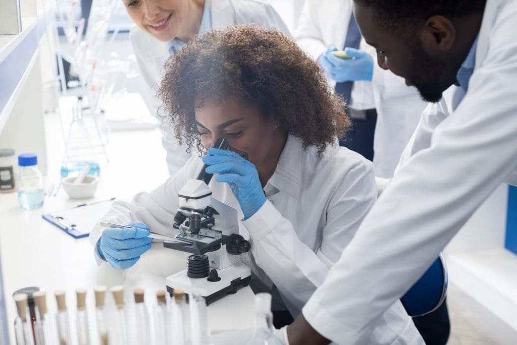 Three scientists talking while one is using a microscope