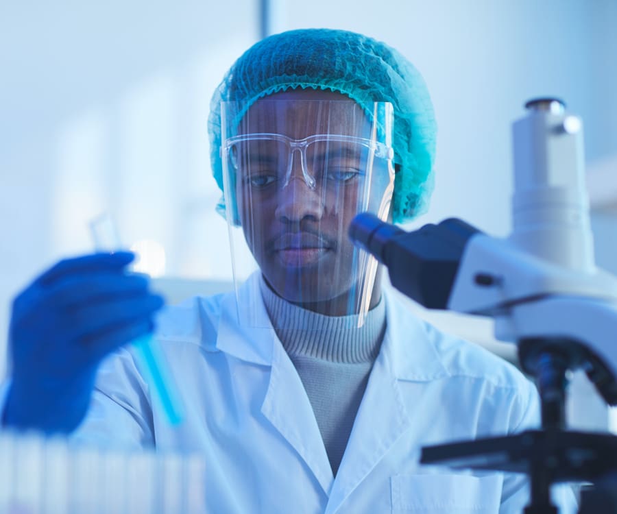 Scientist wearing face shield in front of a microscope while holding a test tube